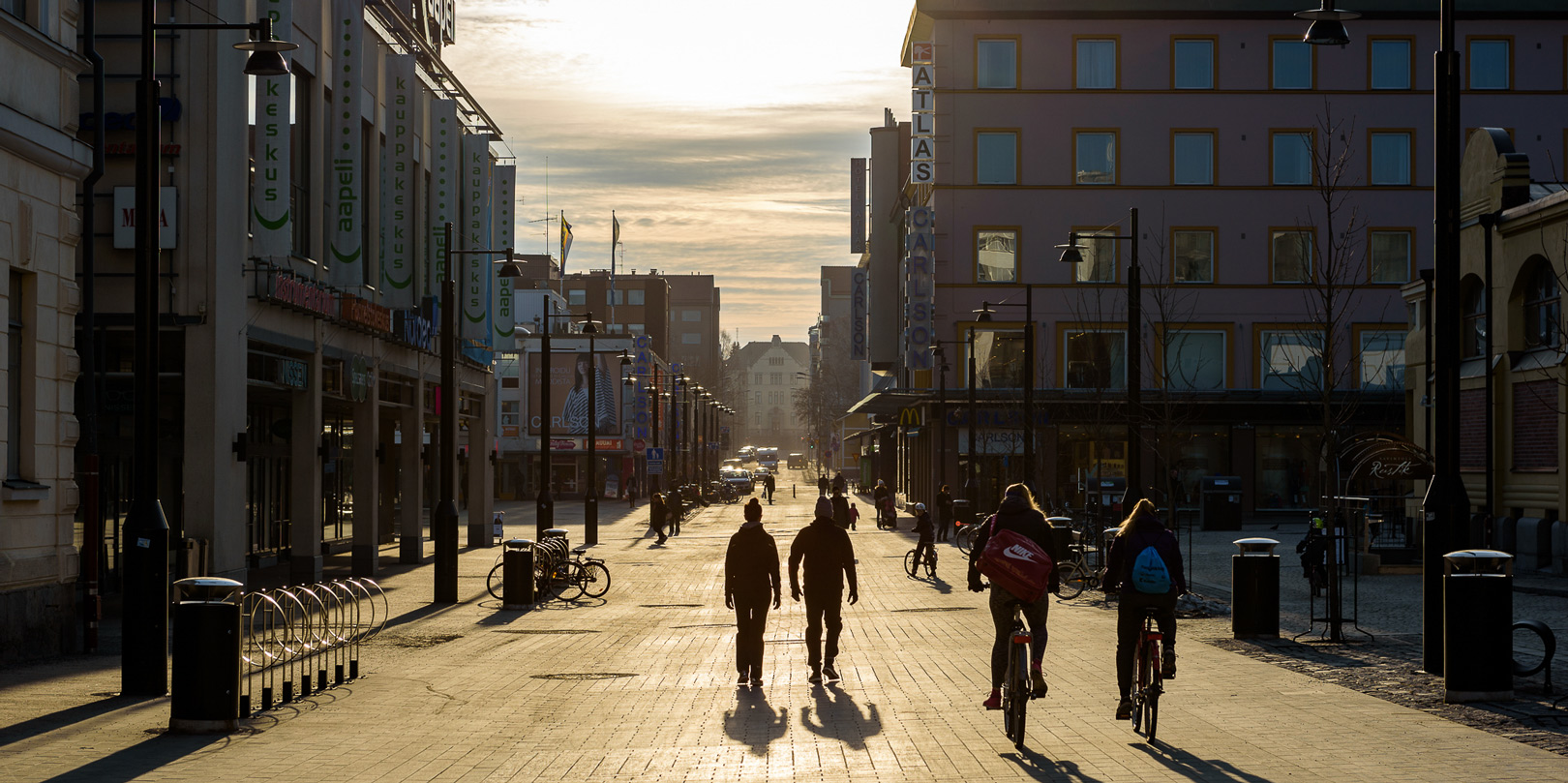 Ihmisiä kävelemässä ja pyöräilemässä Kuopion torilla auringon paistaessa. People walking and cycling in Kuopio Market Square as the sun shines.
