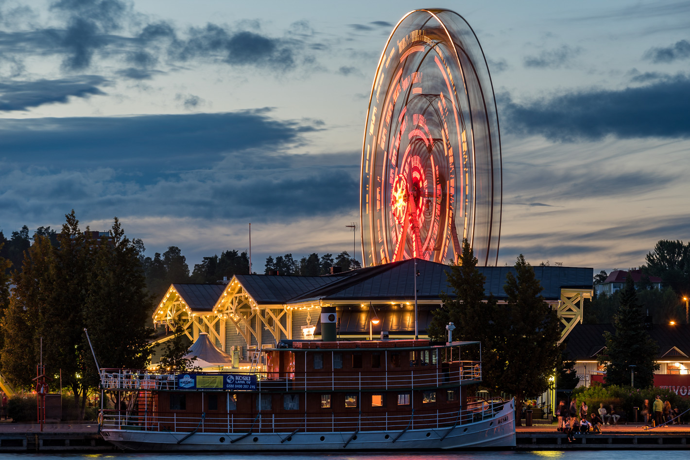 Iltakuva Kuopion satamasta, jossa etualalla on Koski-laiva ja taustalla näkyy valaistu Wanha Satama sekä maailmanpyörä. An evening view of the Port of Kuopio, with the Koski ferry in the foreground and the illuminated Wanha Satama and Ferris wheel visible in the background.