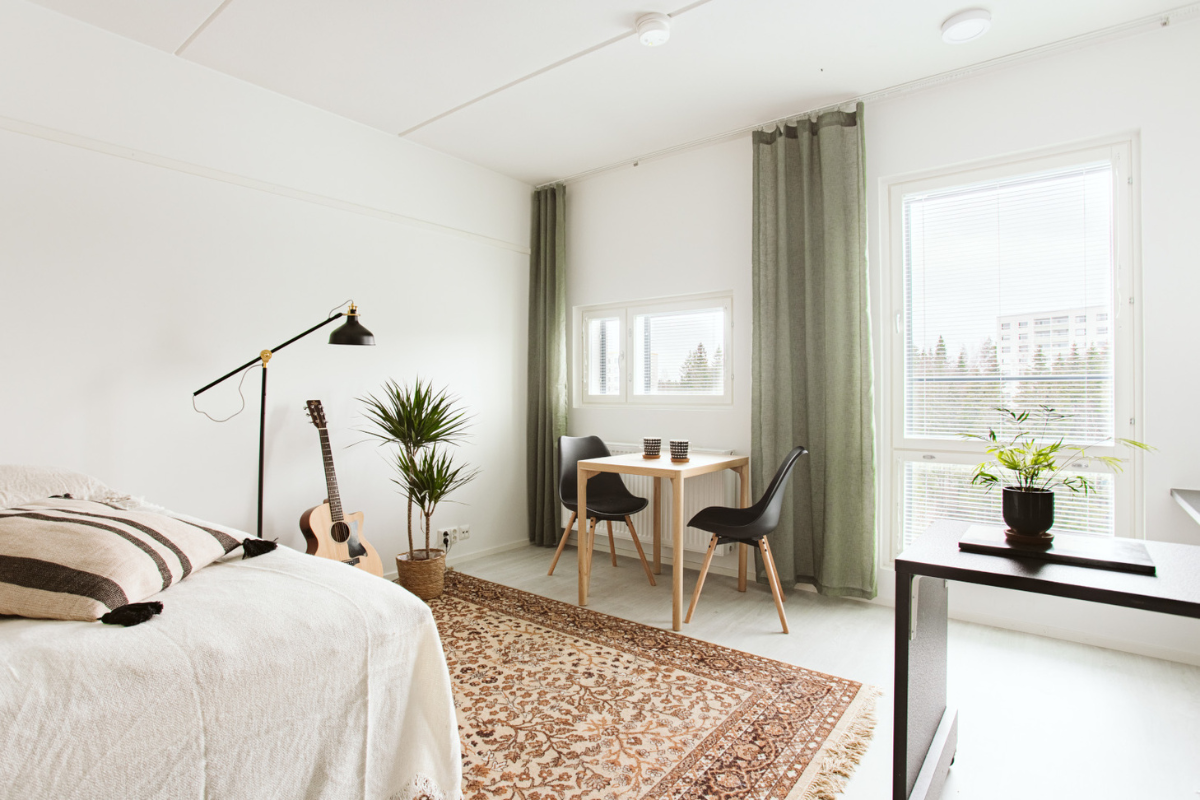 Ahkiotie 8 asunto, jossa näkyy vaalea päiväpeitto sängyn päällä, musta jalkalamppu lattialla, viherkasvi, puinen ruokapöytä ja mustat tuolit, sekä vihreät verhot ja lattialla ruskea matto.