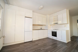 Haapaniementie 24 kolmion vaalea keittiö, josta aukeaa olohuone. Lattia on tummaa puunsävyistä laminaattia. Haapaniementie 24, a bright kitchen opening onto the living room. The floor is dark wood-colored laminate.