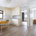 Katiskaniementie 6 kalustetun soluasunnon keittiö, jossa on pöytä ja tuolit. Oikealle aukeaa kaksi kalustettua huonetta. Asunnossa on puunsävyiset laminaattilattiat. Kitchen in a furnished shared apartment at Katiskaniementie 6, with a table and chairs. Two furnished rooms open to the right. The apartment has wood-colored laminate flooring.