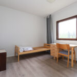 Katiskaniementie 6 kalustetun soluasunnon huone, jossa on ikkunan alla työpöytä ja tuoli, sängynrunko ja patja paketissa sekä lipasto. Lattiat ovat puunsävyistä laminaattia. A furnished room at Katiskaniementie 6, with a desk and chair under the window, a bed frame and mattress in a package, and a chest of drawers. The floors are wood-colored laminate.