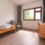 Katiskaniementie 6 kalustetun soluasunnon huone, jossa on ikkunan alla työpöytä ja tuoli, sängynrunko ja patja paketissa sekä korkea hyllykkö ja nojatuoli. Lattiat ovat puunsävyistä laminaattia. A furnished room at Katiskaniementie 6, with a desk and chair under the window, a bed frame and mattress in a package, and high shelf and armchair. The floors are wood-colored laminate.