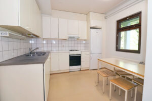 Kuopas Katiskaniementie 6 keittiö, jossa on vaaleat kaapit ja vaalea lattia. Keittiössä on ikkuna ja sen edessä pöytä sekä neljä jakkaraa. The kitchen at Kuopas Katiskaniementie 6 has light-colored cabinets and a light floor. There is a window in the kitchen, with a table and four stools in front of it.