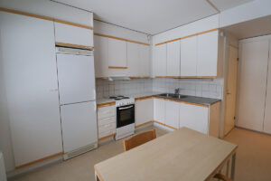 Kuntokuja 2 asunnon keittiö, jossa on vaaleat ovet puunsävyisillä rimoilla. Lattia on vaaleaa muovimattoa ja keittössä on ruokapöytä ja tuoli. Kuntokuja 2 apartment kitchen with light-colored doors and wood-toned slats. The floor is light-colored plastic matting, and the kitchen has a dining table and chair.