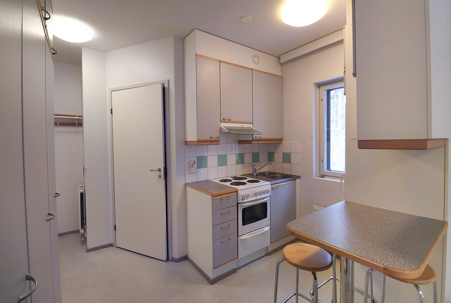 Metsurintie 4 yksiön keittiö ja eteinen. Keittiö on kahdella seinällä ja kaapin vieressä on pieni kiinteä pöytätaso ja jakkarat. Keskellä keittiön seinää on ikkuna. Vasemmalla on vaatekaapit, eteinen ja käynti kylpyhuoneeseen. Lattiat ovat harmaat. Kitchen and hallway of the studio apartment at Metsurintie 4. The kitchen spans two walls, and next to the cabinets there is a small built-in countertop with stools. There is a window in the middle of the kitchen wall. To the left are wardrobes, the hallway, and the entrance to the bathroom. The floors are gray.
