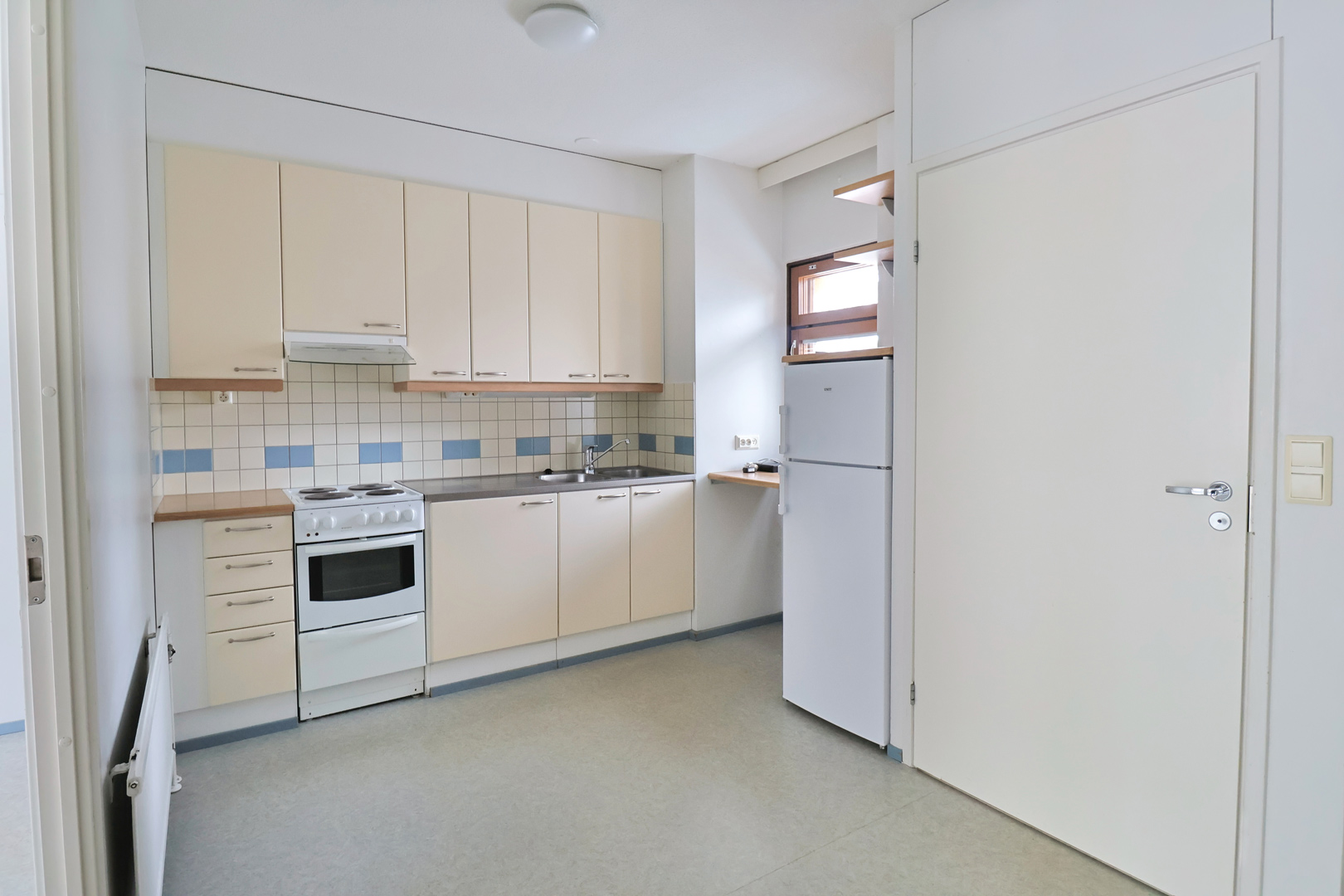 Kuopas Myllärintie 57 asunnon keittiö. Keittiön ovet ovat vaaleat ja nurkassa oven vieressä on ikkuna sekä jääkaappi ja mikro. Lattiat ovat harmaat. Kitchen of the Kuopas Myllärintie 57 apartment. The kitchen cabinets are light-colored, and in the corner next to the door there is a window, a fridge, and a microwave. The floors are grey.