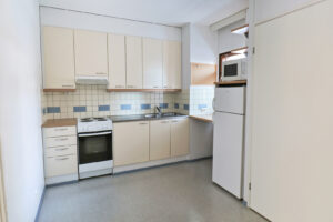 Kuopas Myllärintie 57 asunnon keittiö. Keittiön ovet ovat vaaleat ja nurkassa oven vieressä on ikkuna sekä jääkaappi ja mikro. Lattiat ovat harmaat. Kitchen of the Kuopas Myllärintie 57 apartment. The kitchen cabinets are light-colored, and in the corner next to the door there is a window, a fridge, and a microwave. The floors are grey.