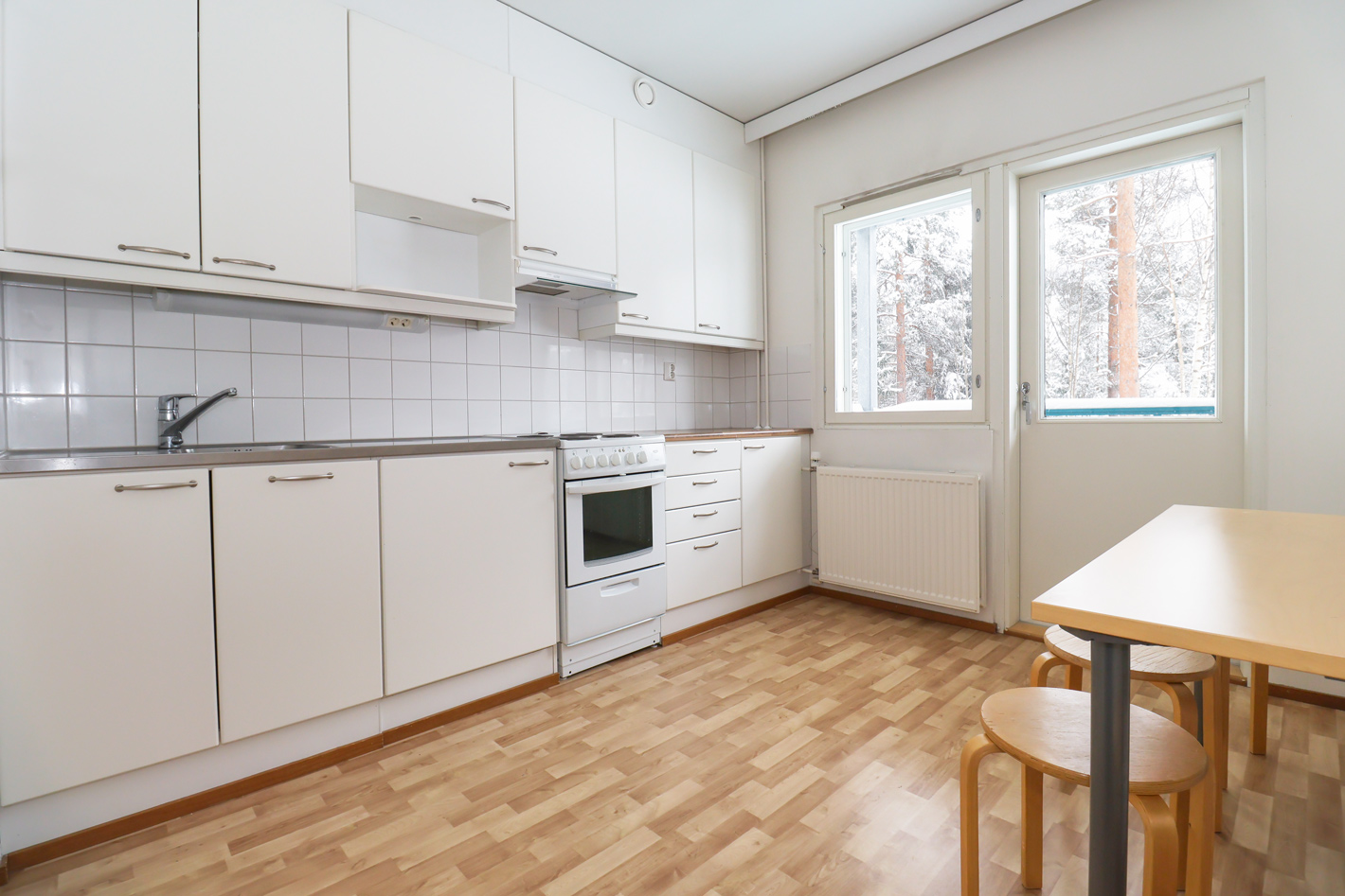 Neulamäentie 3 asunnon valkoinen keittiö. Oikealla on ikkuna ja parvekkeen ovi sekä puinen pöytä ja jakkarat. Lattia on puun värinen. The white kitchen in the apartment at Neulamäentie 3. On the right are a window and the balcony door, as well as a wooden table and stools. The floor is wood-colored.