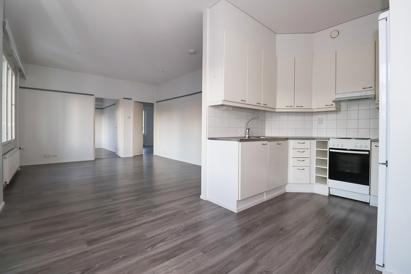 Pyöräkatu 7 vaalea keittiö oikealla ja vasemmalla aukeaa näkymä olohuoneeseen. Olohuoneesta pääsee kahteen makuuhuoneesen. Lattiat ovat tumman puun sävyistä laminaattia. The light-colored kitchen is on the right at Pyöräkatu 7, and the living room is visible to the left. The living room leads to two bedrooms. The floors are dark wood-toned laminate.