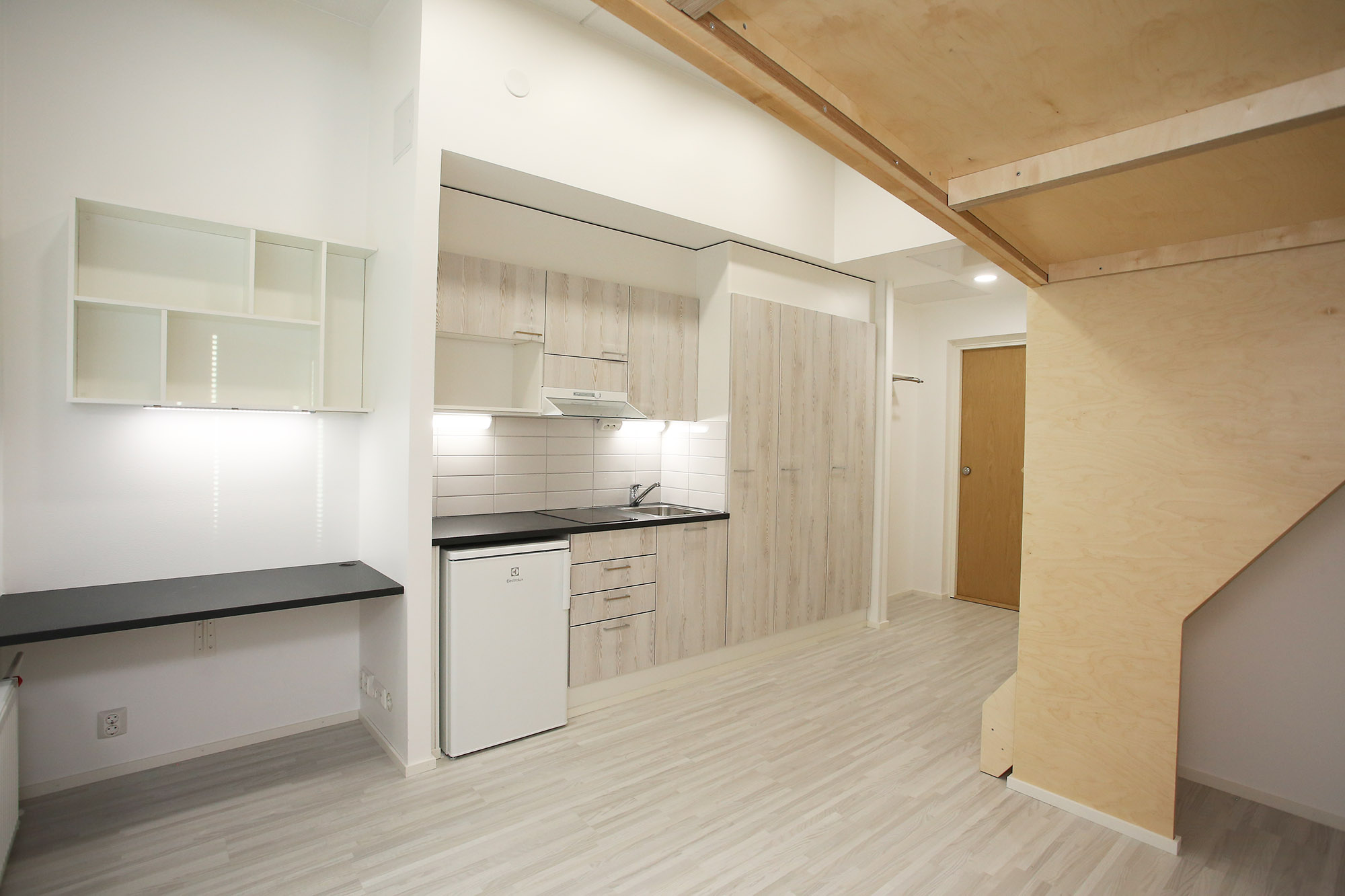 Samoilijantie 6 asunto, jossa kuva otettu nukkumaparven alta. Ikkunan edessä ovat kiinteä seinähylly sekä tumma työpöytätaso ja sen vieressä keittiö ja korkeita kaappeja, joissa on vaalean puun sävyiset kaapin ovet. Lattia on puunsävyistä laminaattia. Samoilijantie 6 apartment, with the photo taken from under the loft bed. In front of the window are a built-in wall shelf and a dark desk; next to it is the kitchen and tall cabinets with light wood-toned doors. The floor is wood-toned laminate.