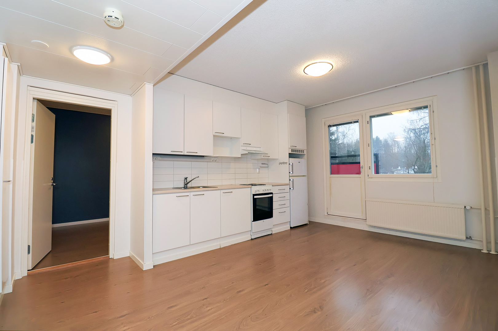 Särkiniementie 18 avaran asunnon vaalea keittiö, jonka oikealla puolen on ranskalainen parveke ja ikkuna, ja vasemmalla puolen ovi makuuhuoneeseen. Lattiat ovat puunväristä laminaattia. Light-colored kitchen in the spacious apartment at Särkiniementie 18, with a French balcony and window on the right and a door to the bedroom on the left. The floors are wood-colored laminate.