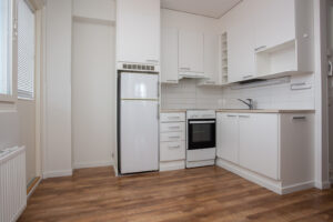Kuopas Särkiniementie 18 asunto, jossa on valkoinen pieni kulmakeittiö, puunväriset lattiat ja vasemmalla ikkuna sekä parvekkeen ovi. Kuopas Särkiniementie 18 apartment, which has a small white corner kitchen, wood-colored floors and on the left there's a window and a balcony door.