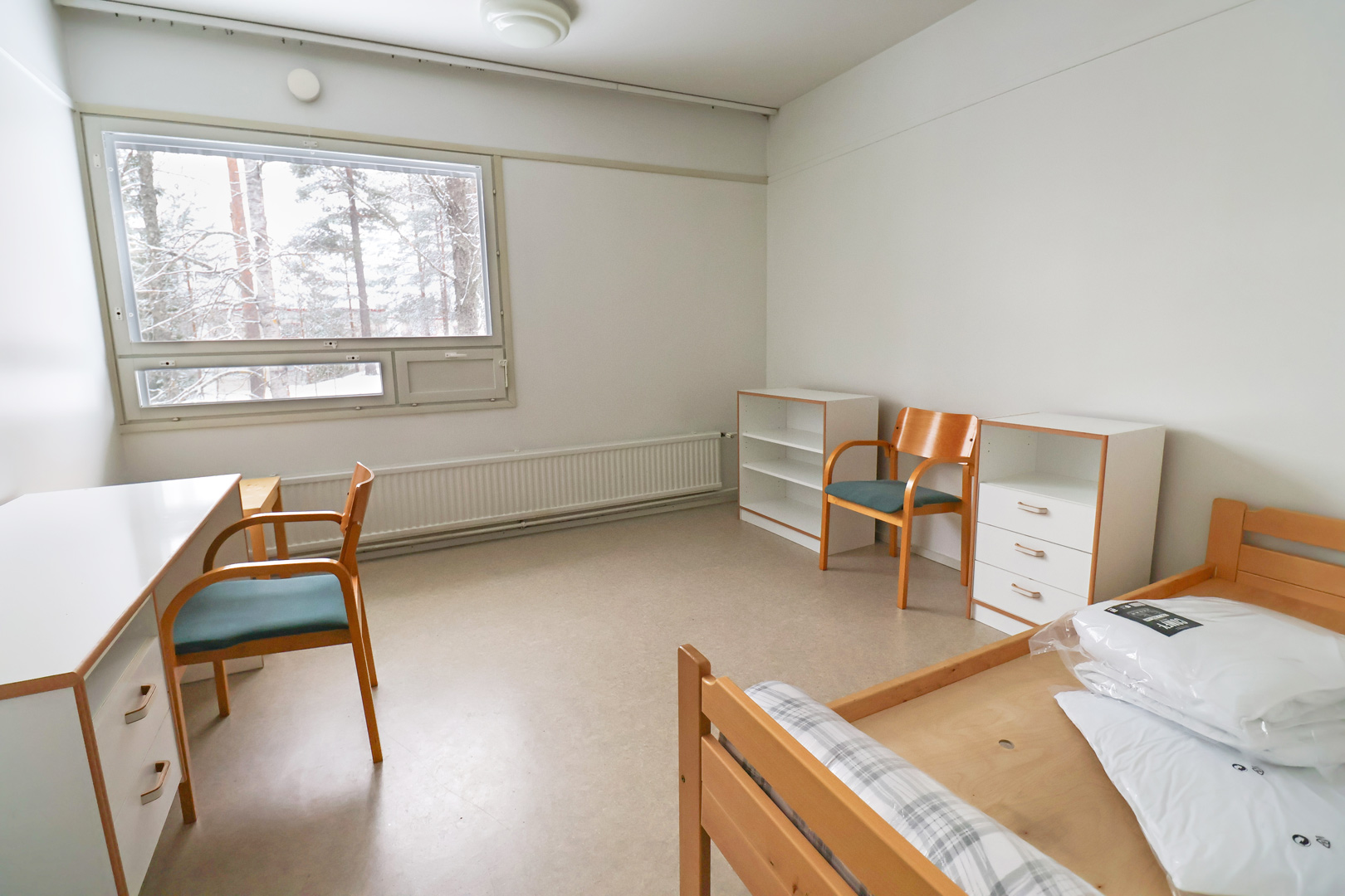 Kuopas Särkiniementie 30 kalustettu miniyksiö. Oikealla on puunvärinen sängynrunko ja kaksi lipastoa sekä tuoli. Vasemmalla ikkunan edessä on valkoinen työpöytä ja puunvärinen tuoli. Kuopas Särkiniementie 30 furnished mini studio. On the right there is a wooden bed frame and two chests of drawers and a chair. On the left in front of the window is a white desk and a wooden chair.