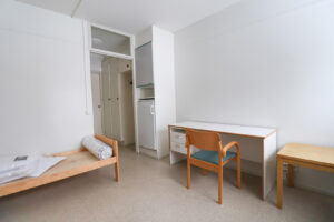 Kuopas Särkiniementie 30 kalustettu miniyksiö. Vasemmalla on puunvärinen sängynrunko ja oikealla valkoinen työpöytä ja puunvärinen tuoli. Taustalla näkyy eteinen. Kuopas Särkiniementie 30 furnished mini studio apartment. On the left is a wood-colored bed frame and on the right a white desk and wood-colored chair. The hallway is visible in the background.