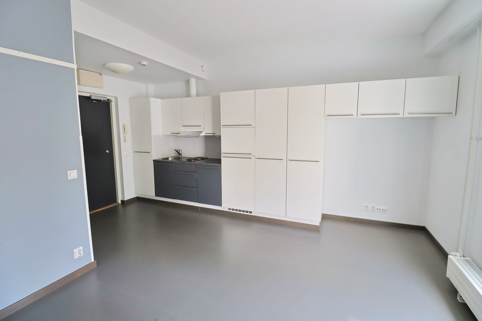 Satamakatu 26 asunnon yksiö, jossa ulko-oven vieressä on keittiö. Keittiöstä jatkuu korkeita kaappeja ikkunaseinään saakka. Ikkunan vieressä on vain ylhäällä korkeita kaappeja, jotta lattialla voi pitää esimerkiksi työpöytää. Lattiat ovat tummanharmaat. A studio apartment at Satamakatu 26, with the kitchen located next to the front door. Tall cabinets run from the kitchen all the way to the window wall. Next to the window, there are only tall cabinets at the top, leaving space on the floor for a desk, for example. The floors are dark gray.